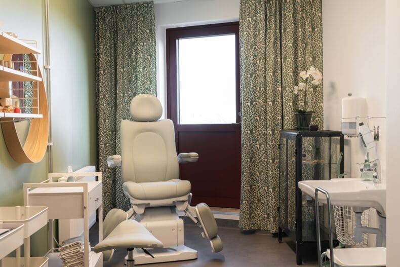 Behandlingsrum. Ljust behandlingsrum med grönmönstrade gardiner, bekväm behandlingsstol, handfat och hyllor för utrustning.