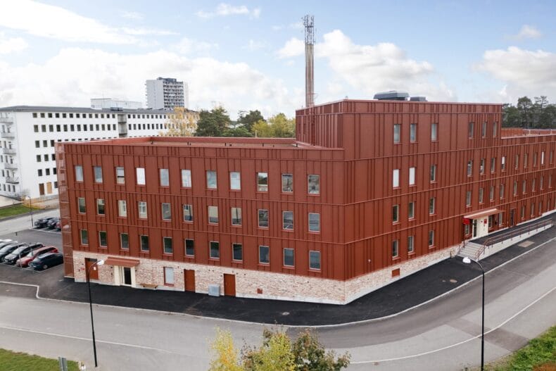 Närbild på byggnadens fasad. Tegelröd byggnad i flera våningar med stora fönster, stenklädd sockel och parkeringsplats utanför.