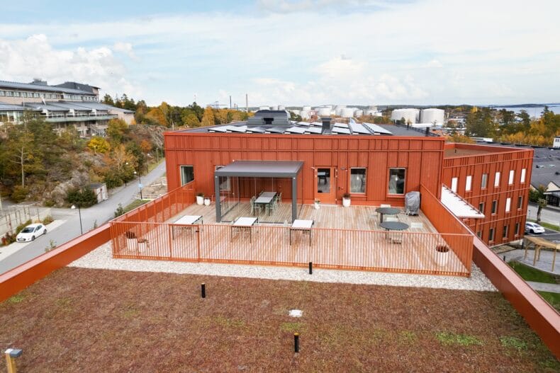 Takterrass med sittplatser. Stor takterrass med bord, stolar och ett glastak över en del av ytan, omgiven av räcken och med utsikt över staden och havet.