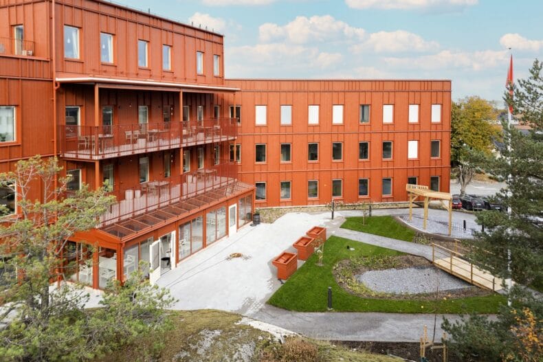 Innergård med grönska. Byggnadens baksida med uteplats, planteringslådor, gångvägar och träd, omgiven av tegelröd fasad och balkonger.