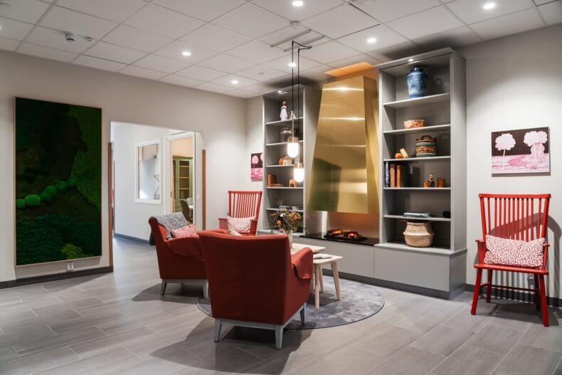 Inomhusmiljö med sittgrupp. Ett mysigt sällskapsrum med röda fåtöljer, en guldig öppen spis, bokhyllor och varm belysning som skapar en hemtrevlig känsla.