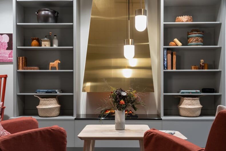 Närbild på eldstad och bokhyllor. Guldig eldstad mellan två bokhyllor med böcker, keramik och inredningsdetaljer, med blommor i vas på ett litet bord framför.