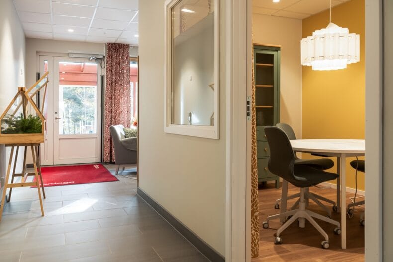 Korridor med mötesrum. Ljus korridor med stolar, växter och ett mindre mötesrum med gula väggar och rund sittgrupp synligt genom dörröppningen.