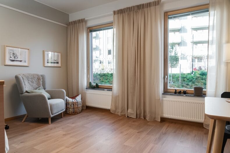 Ljust rum med fåtölj. Hemtrevligt rum med stora fönster, ljusa gardiner och en fåtölj med filt, kudde och korg med textilier. boendes lägenhet