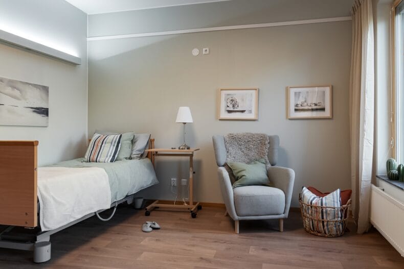 Sovrum med säng och fåtölj. Boenderum med justerbar säng, fåtölj, nattduksbord och tavlor på väggen, inrett i lugna färger.