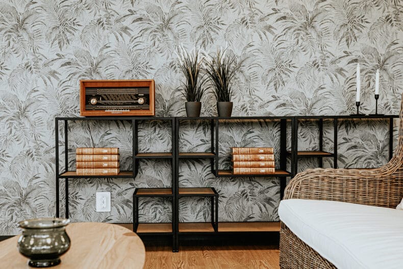 Låg bokhylla med böcker, två gröna växter, ljusstakar och en gammal radio mot mönstrad grå tapet