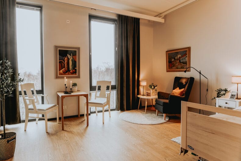 Boendelägenhet med mörk fåtölj och vitt sidobord och lampa i hörnet, kvadratiskt bord i trä med två vita stolar. Mörka gardiner mot stora fönster