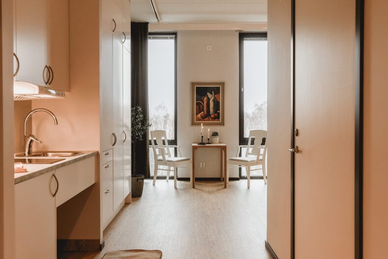 Lägenhet med diskho och kokplatta och vita skåp, litet bord med två vita stolar mellan fönter